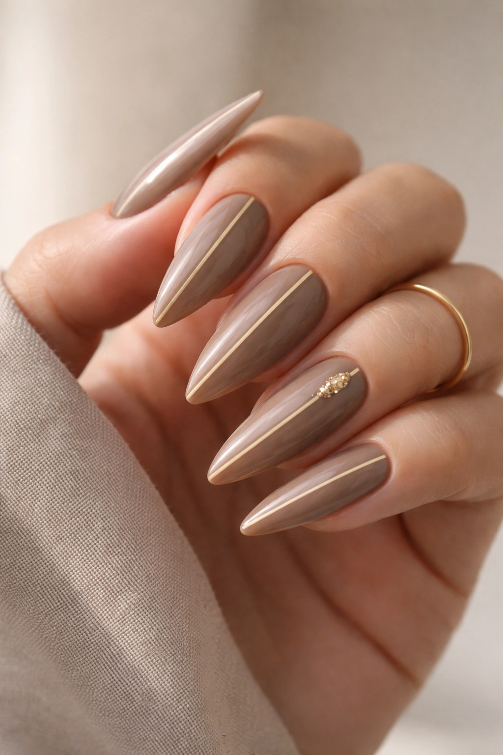Close-up of taupe stiletto nails with a gold centerline accent on a neutral background