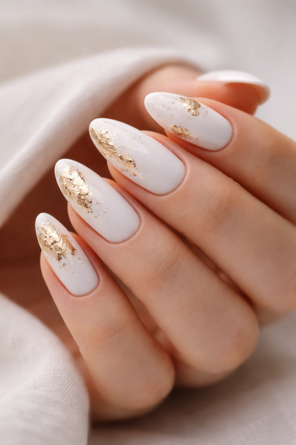 Close-up of white almond nails with gold foil accents on plain background