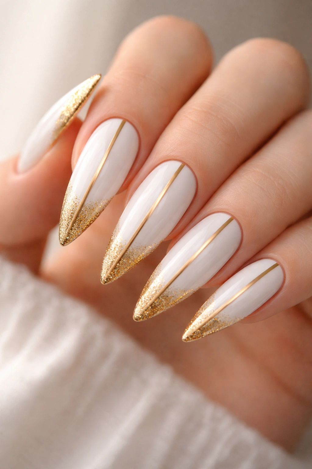 Close-up of white stiletto nails with gold glitter tips and a thin vertical stripe