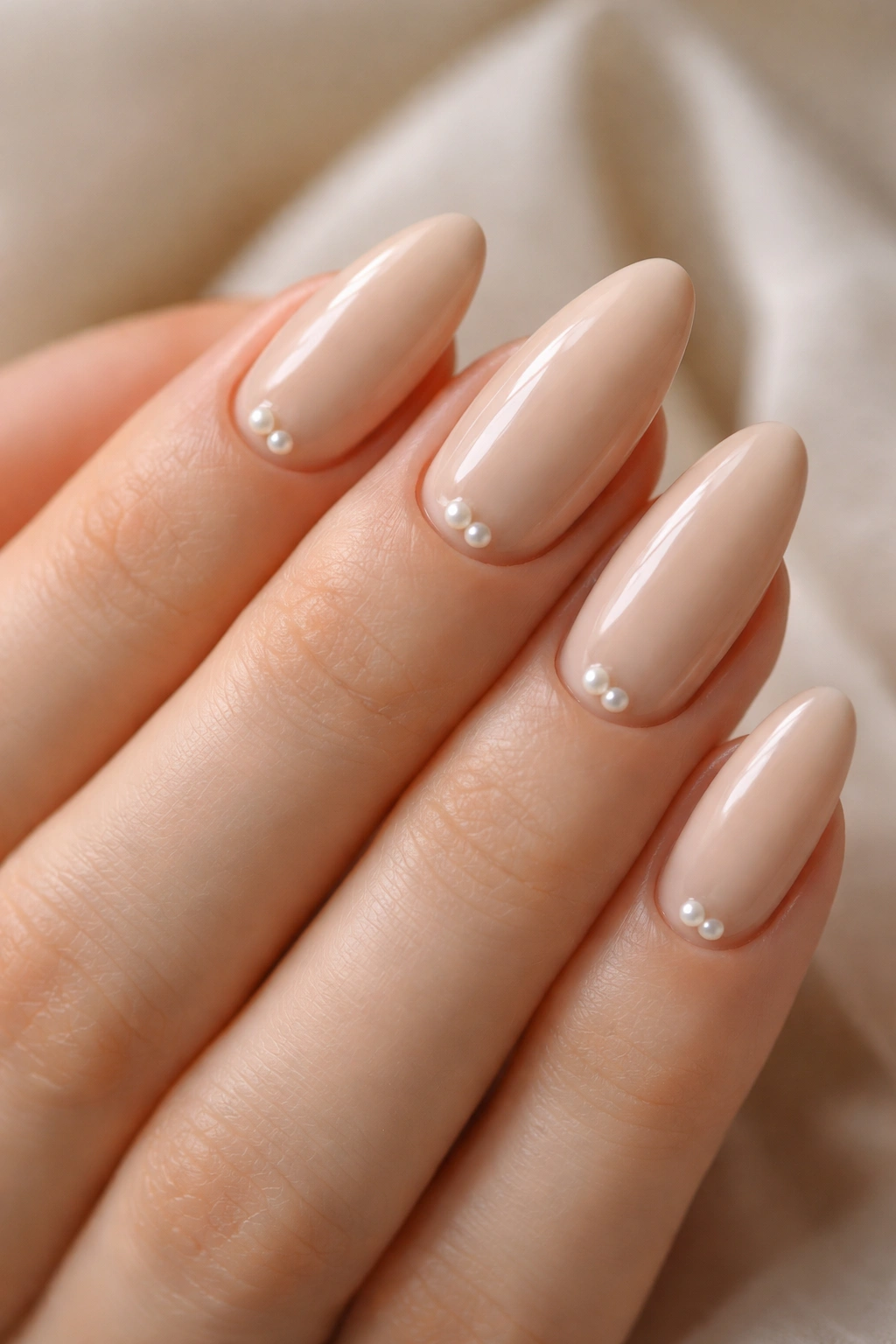 Close-up of beige almond nails with tiny pearl studs on a soft, neutral background
