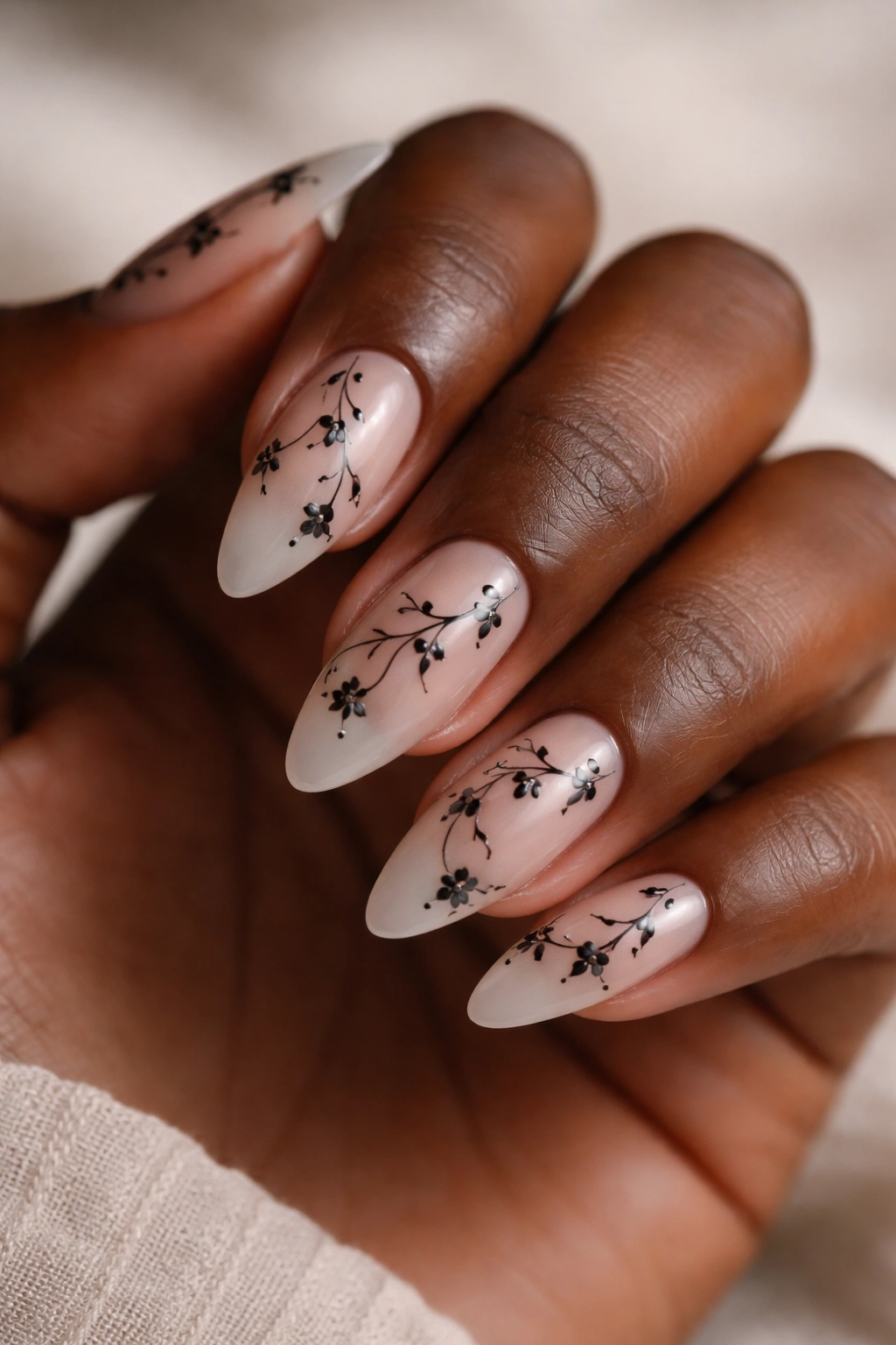 Close-up of almond nails with black floral vine art on deep skin tone, sheer blush base, moody studio lighting