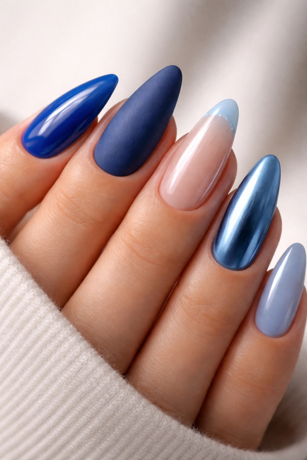Close-up of blue almond nails with mixed finishes on a hand: glossy cobalt, matte navy, chrome, jelly pale blue, and micro-French accents