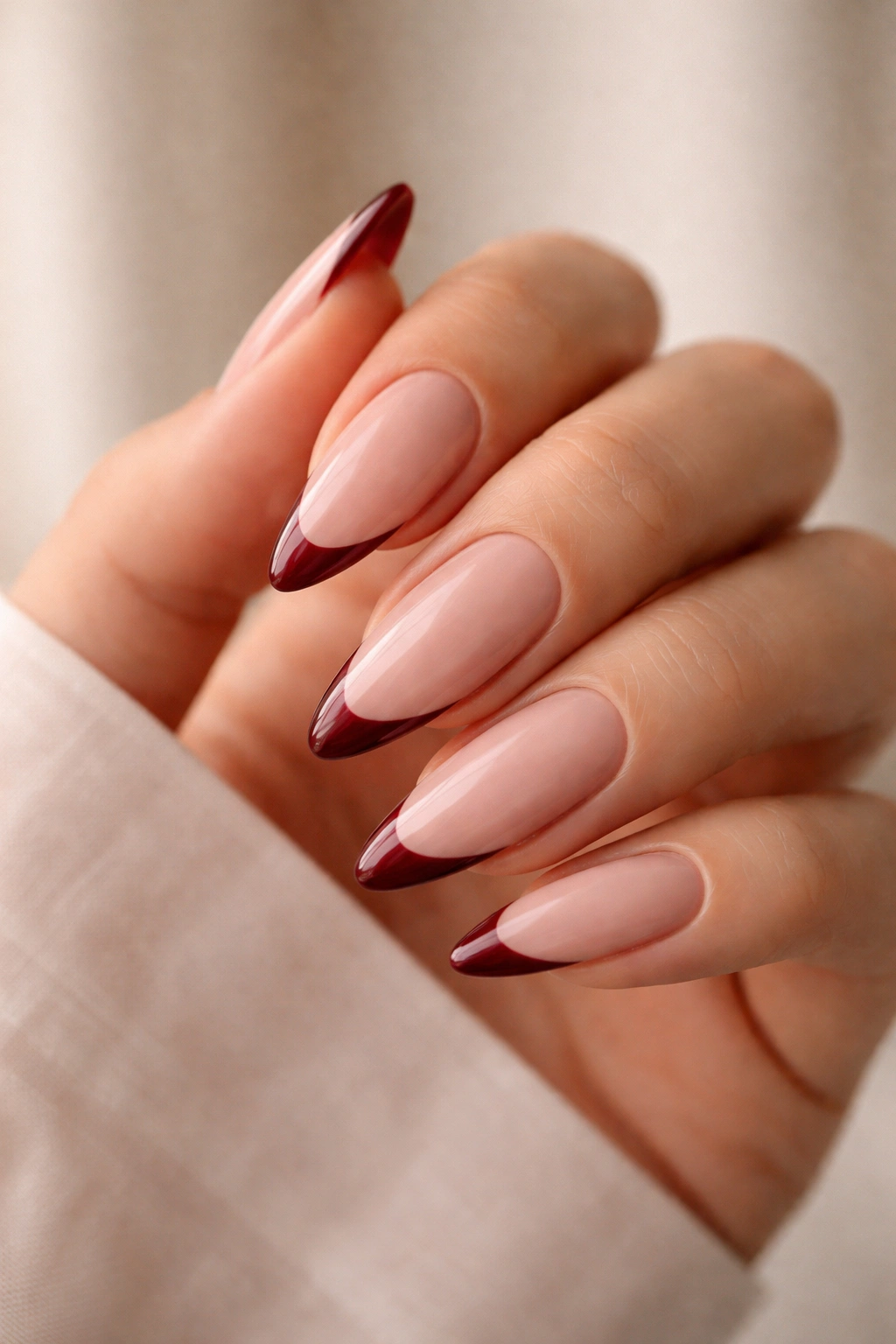 Close-up of a hand with long almond nails featuring a thin burgundy French tip on a nude base, glossy finish.