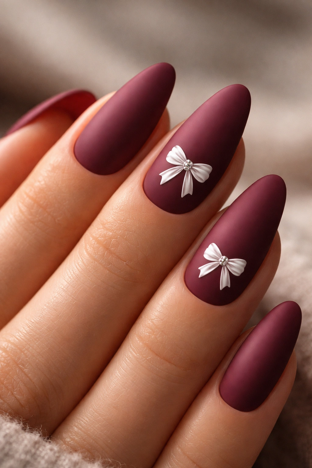 Matte burgundy almond nails with a glossy bow sticker on one nail in a minimal studio setting.