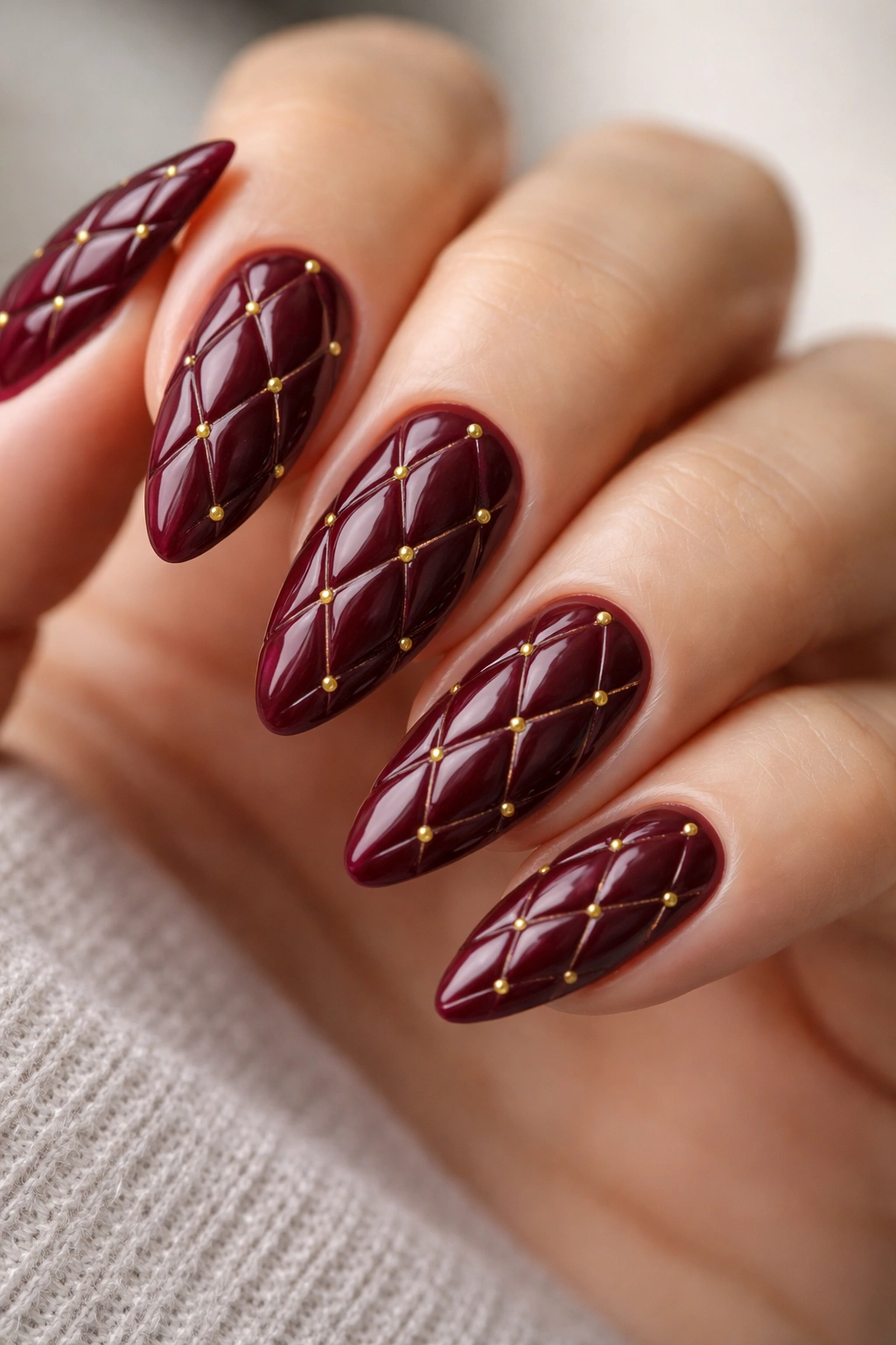 Close-up of burgundy quilted texture nails on almond-shaped fingertips showing raised diamond pattern and tiny gold beads at intersections