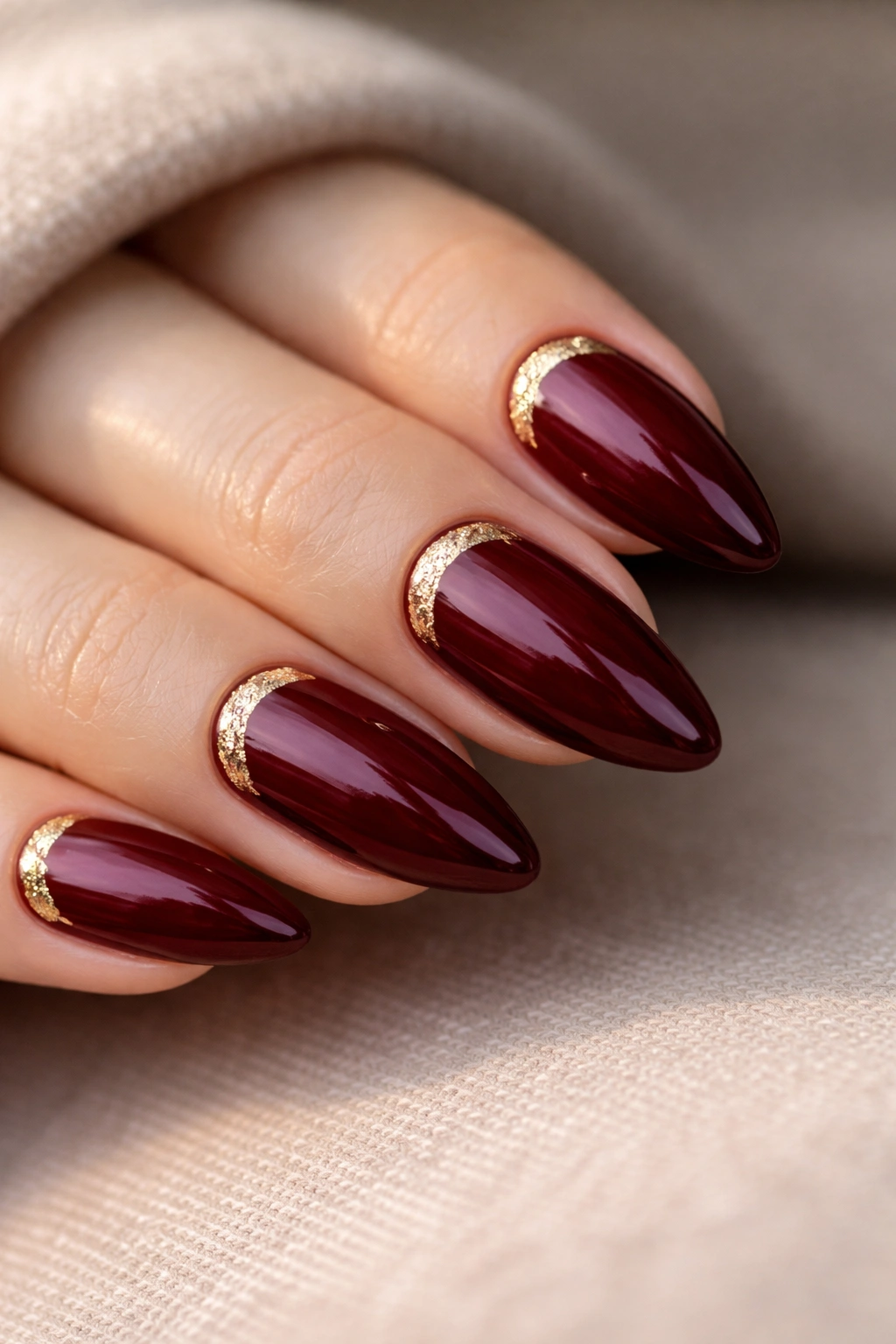 Close-up of dark red almond nails with a slim gold leaf crescent near the cuticle on glossy polish