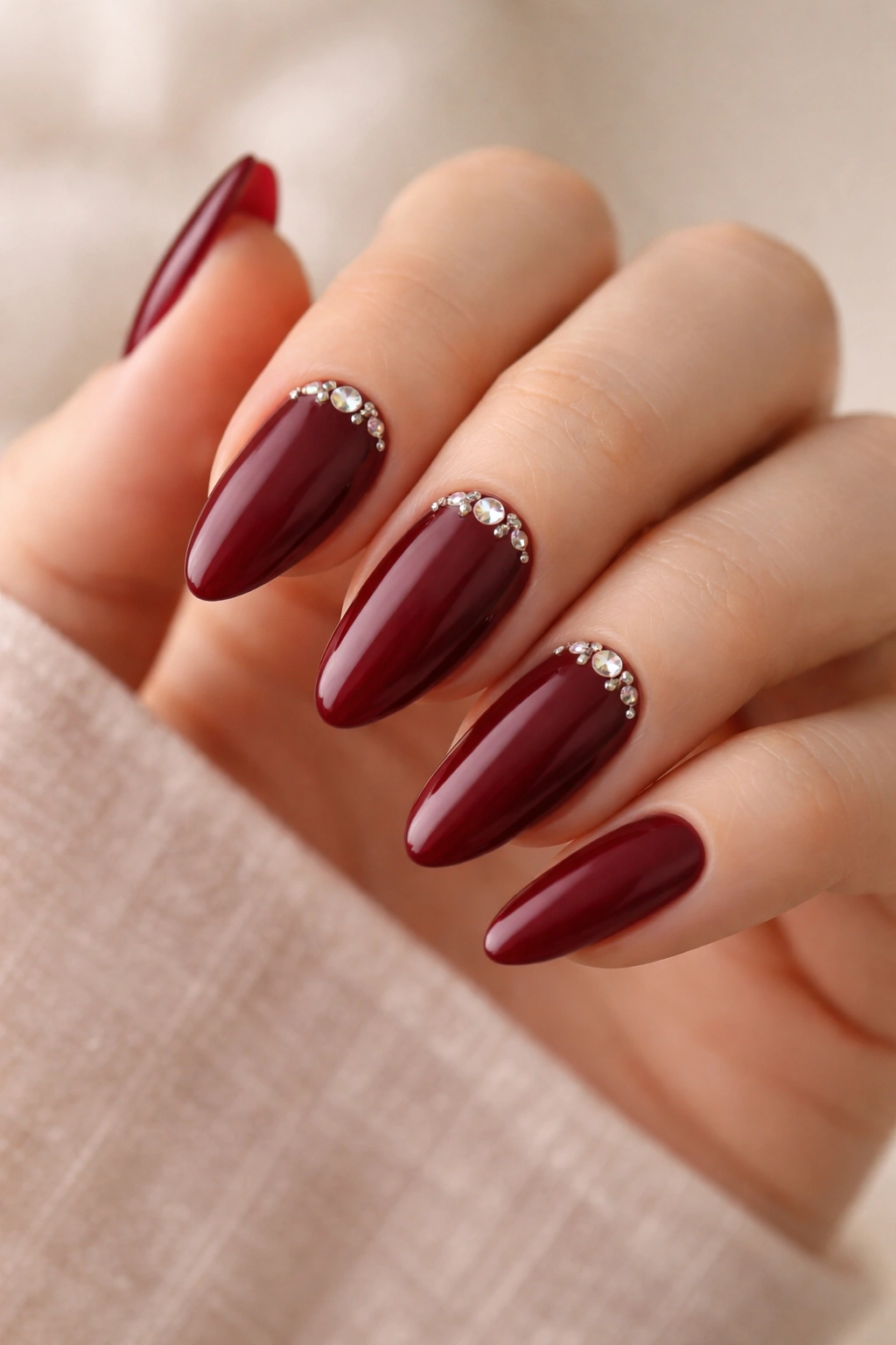 Almond nails in maroon with small crystals near the cuticle on a neutral backdrop