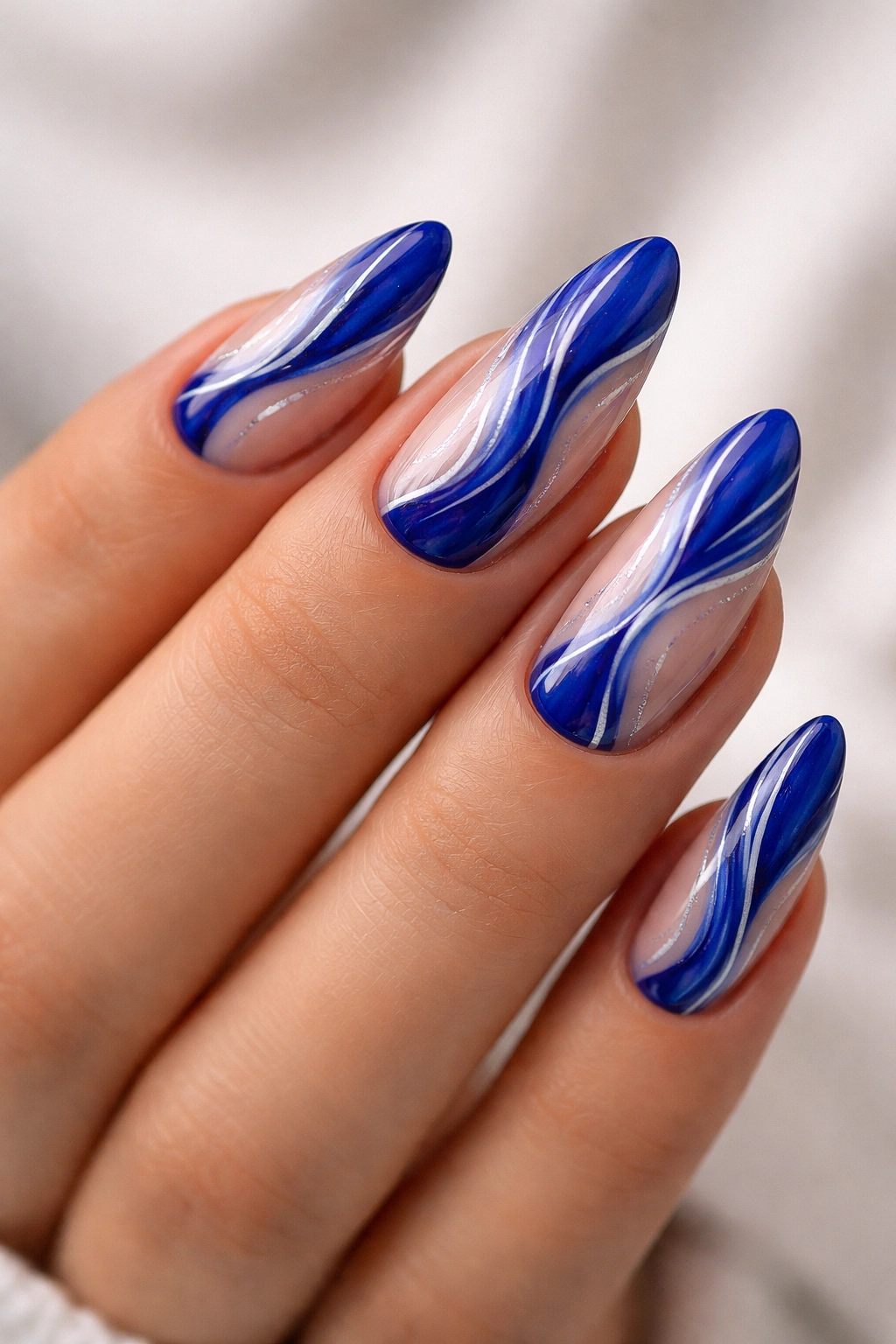 Close-up of almond nail with royal blue abstract wave design on nude base, white and silver accents