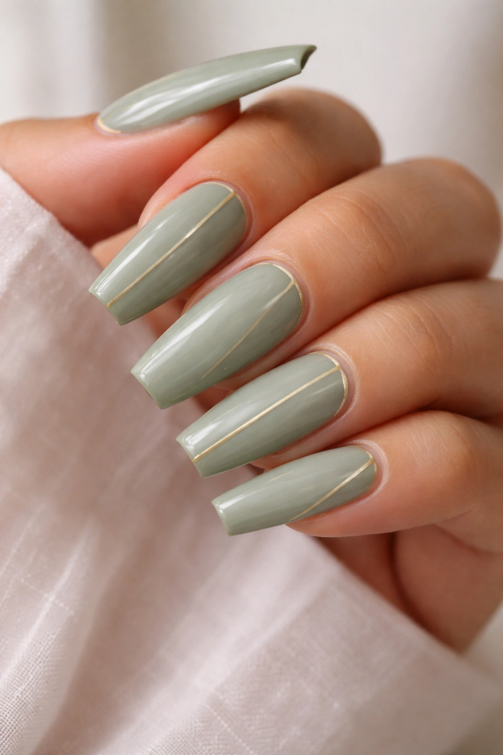 Close-up of sage green coffin nails with slim gold lines on a plain background.