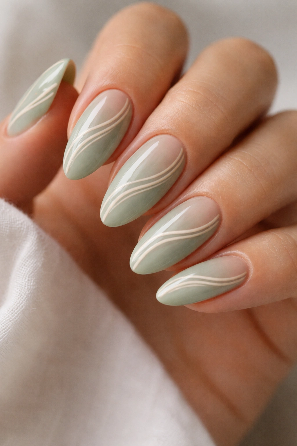 Close-up of almond nails with sage green base and thin white-beige swirls along the curve