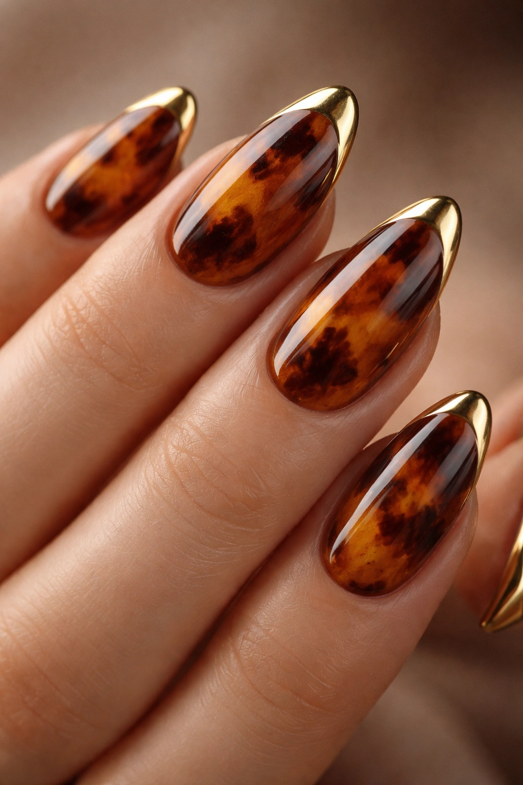 Close-up of tortoiseshell almond nails with gold corner tips on a neutral backdrop