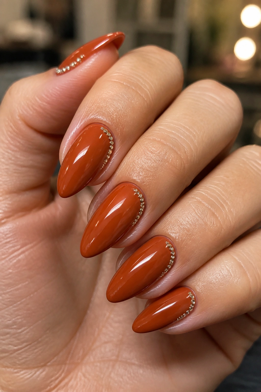Close-up of almond nails in burnished orange with crystal dot accents near the cuticle