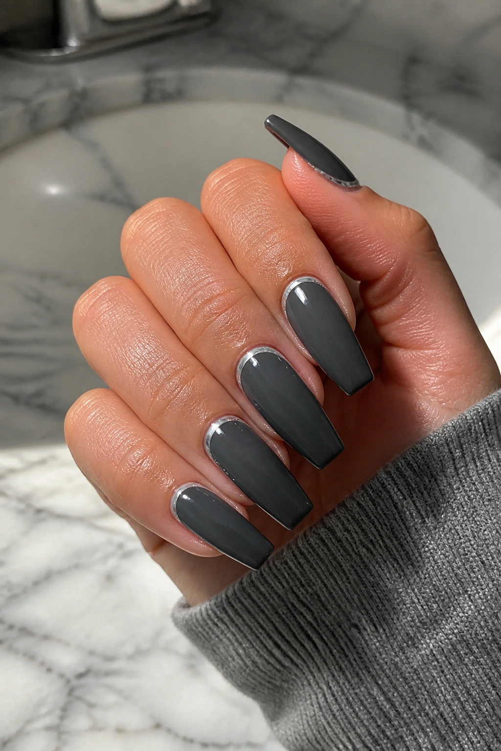 Close-up of charcoal grey coffin nails with a thin silver cuticle line on a pale marble surface