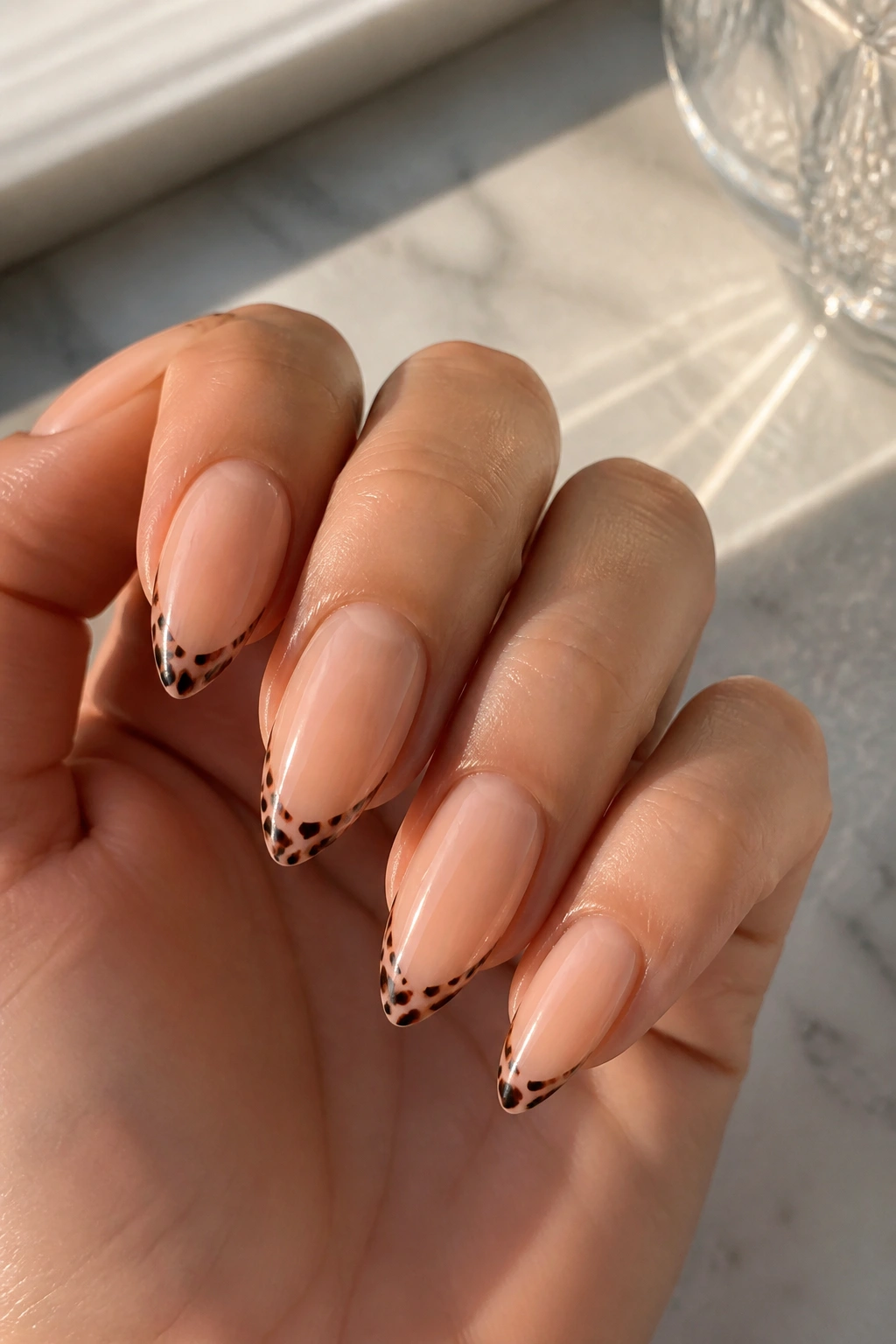Close-up of almond nails with thin cheetah tips on a sheer nude base in warm light