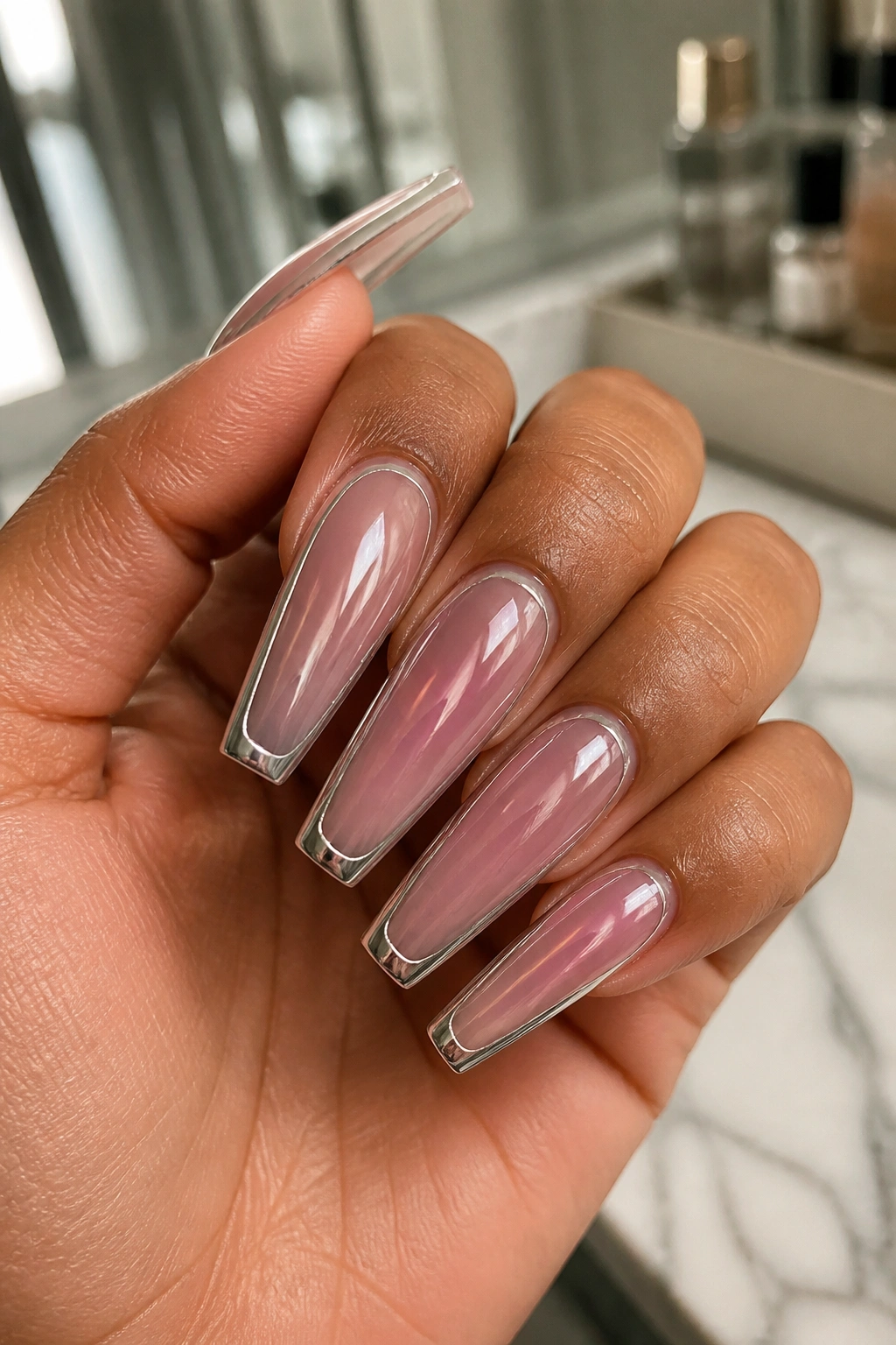 Close-up of coffin nails with chrome edges and frosted pink tips on a real hand in a softly lit bathroom vanity.