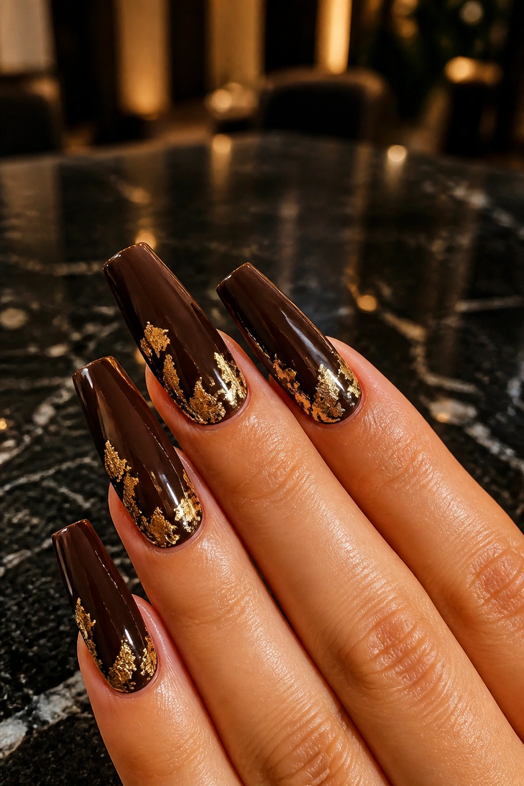 Close-up of hands with Dark Truffle Coffin Nails With Gold Foil featuring deep brown nails and gold foil accents
