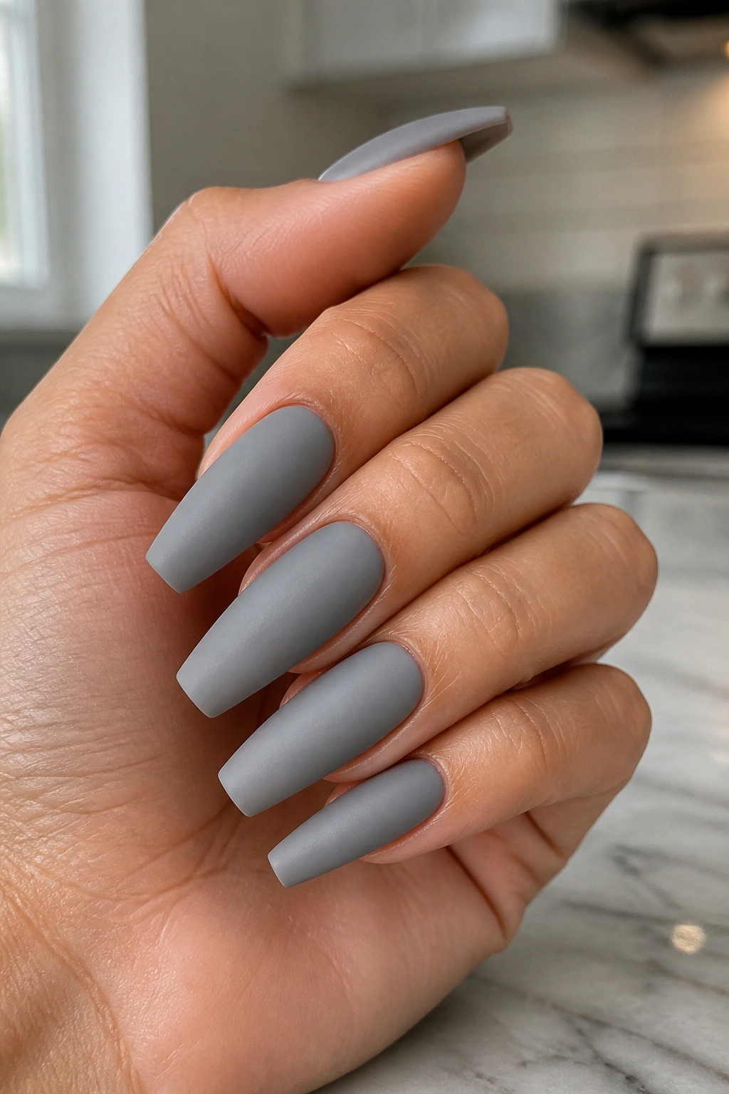 Close-up of a real hand showing matte dove grey coffin nails on a marble counter