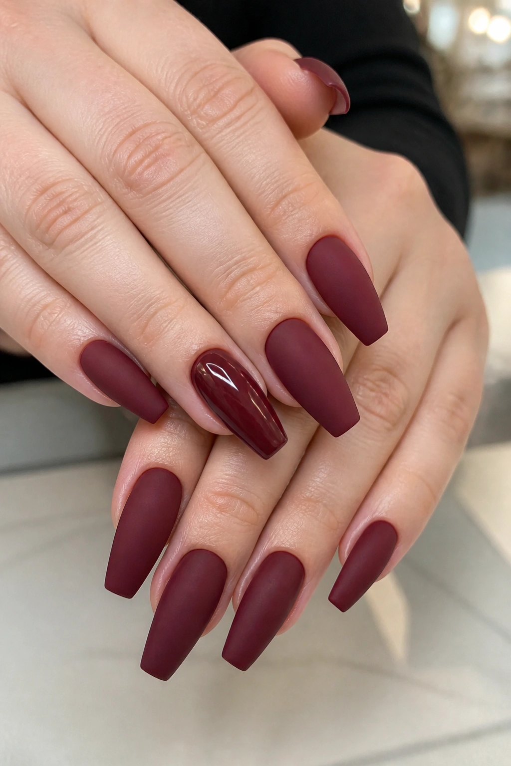 Close-up of hands with matte burgundy nails and a single glossy accent nail