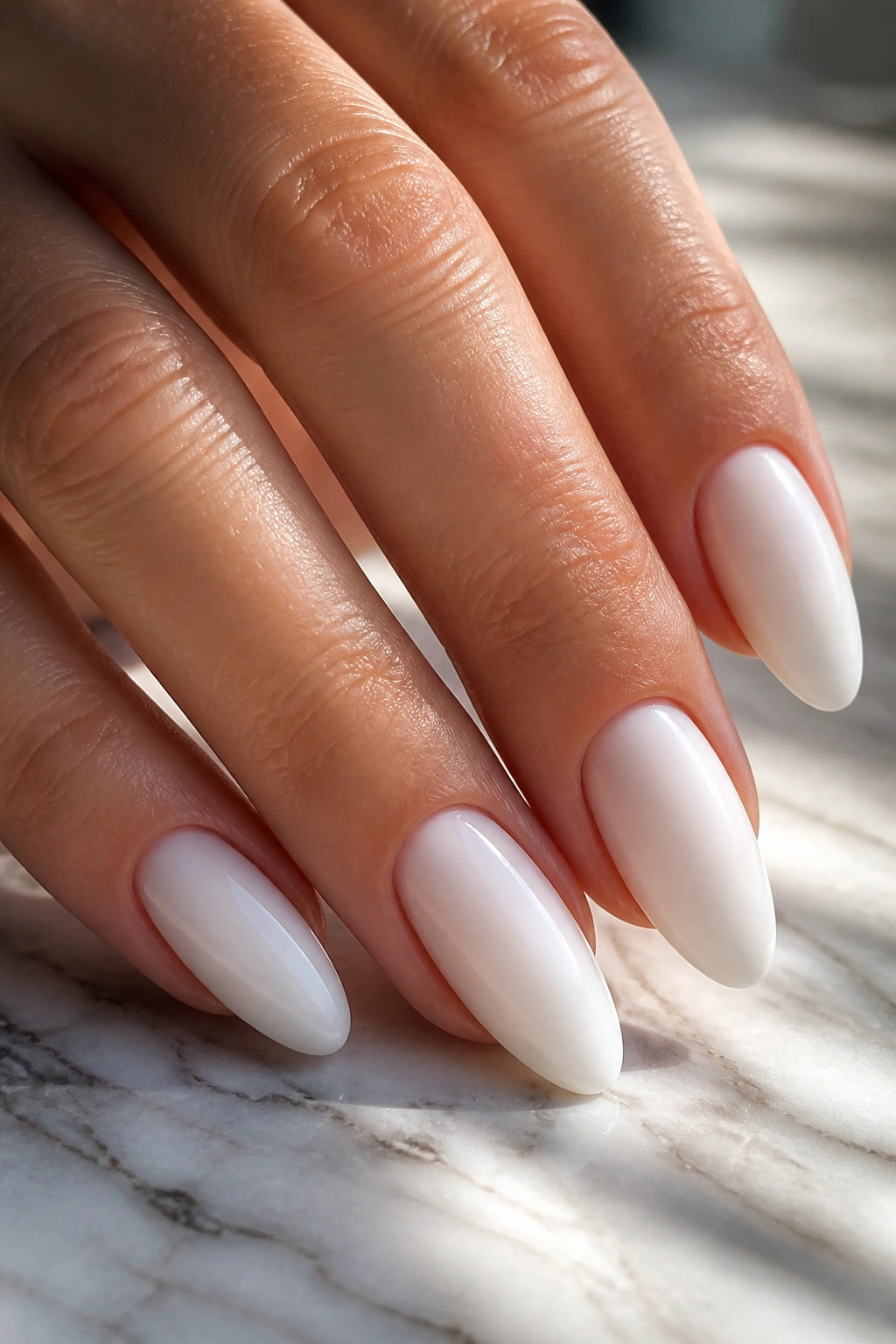 Close-up of a real hand with milky white almond nails on marble surface in warm light