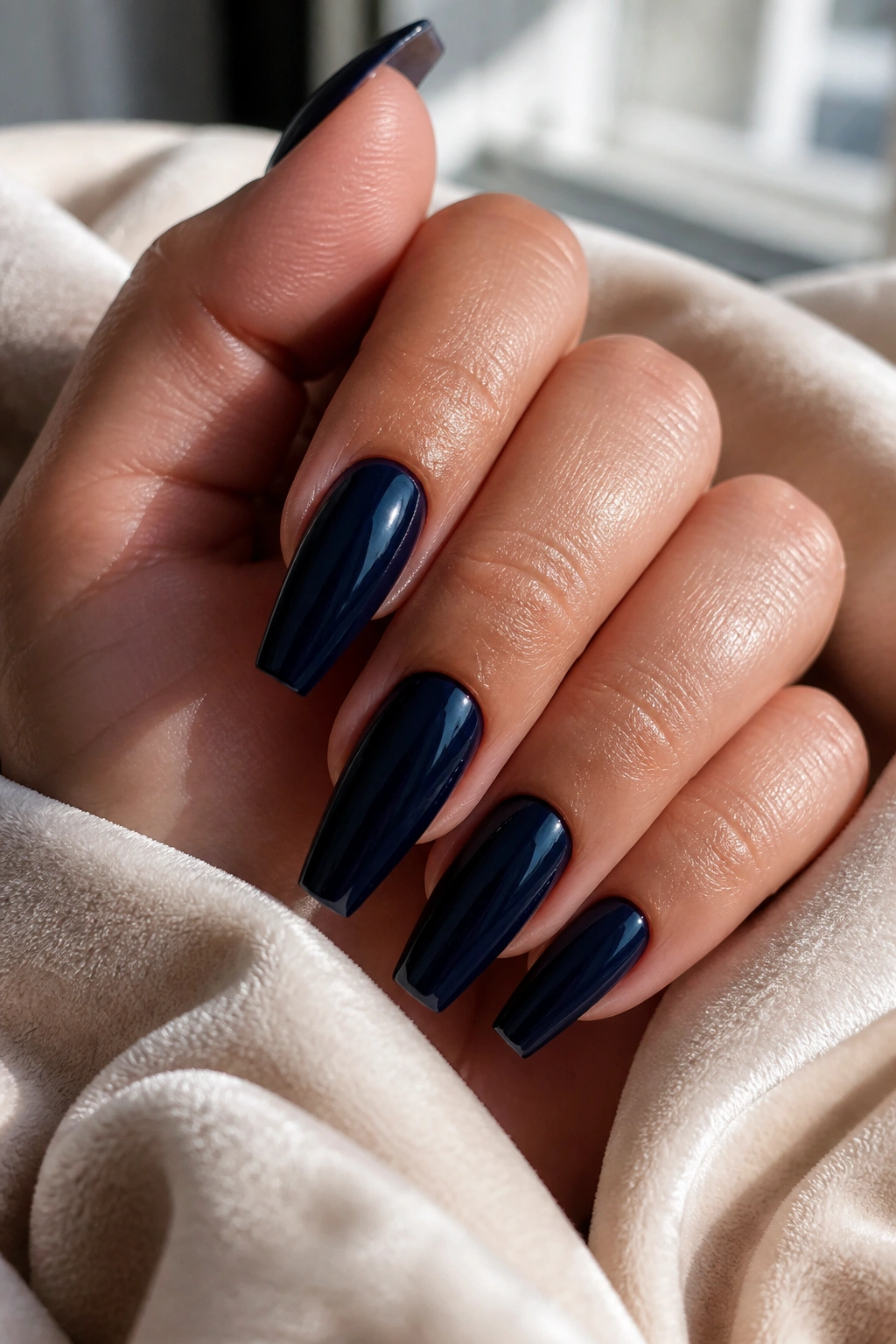 Close-up of a real hand with deep navy coffin nails on cream velvet, glossy finish