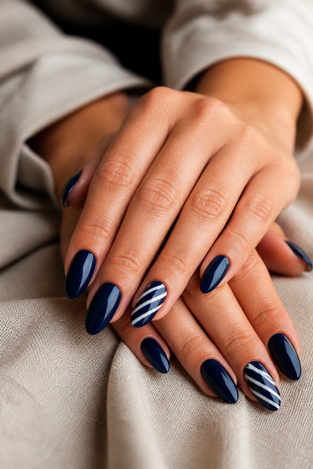 Close-up of almond nails in navy with white stripe accents on a soft linen surface