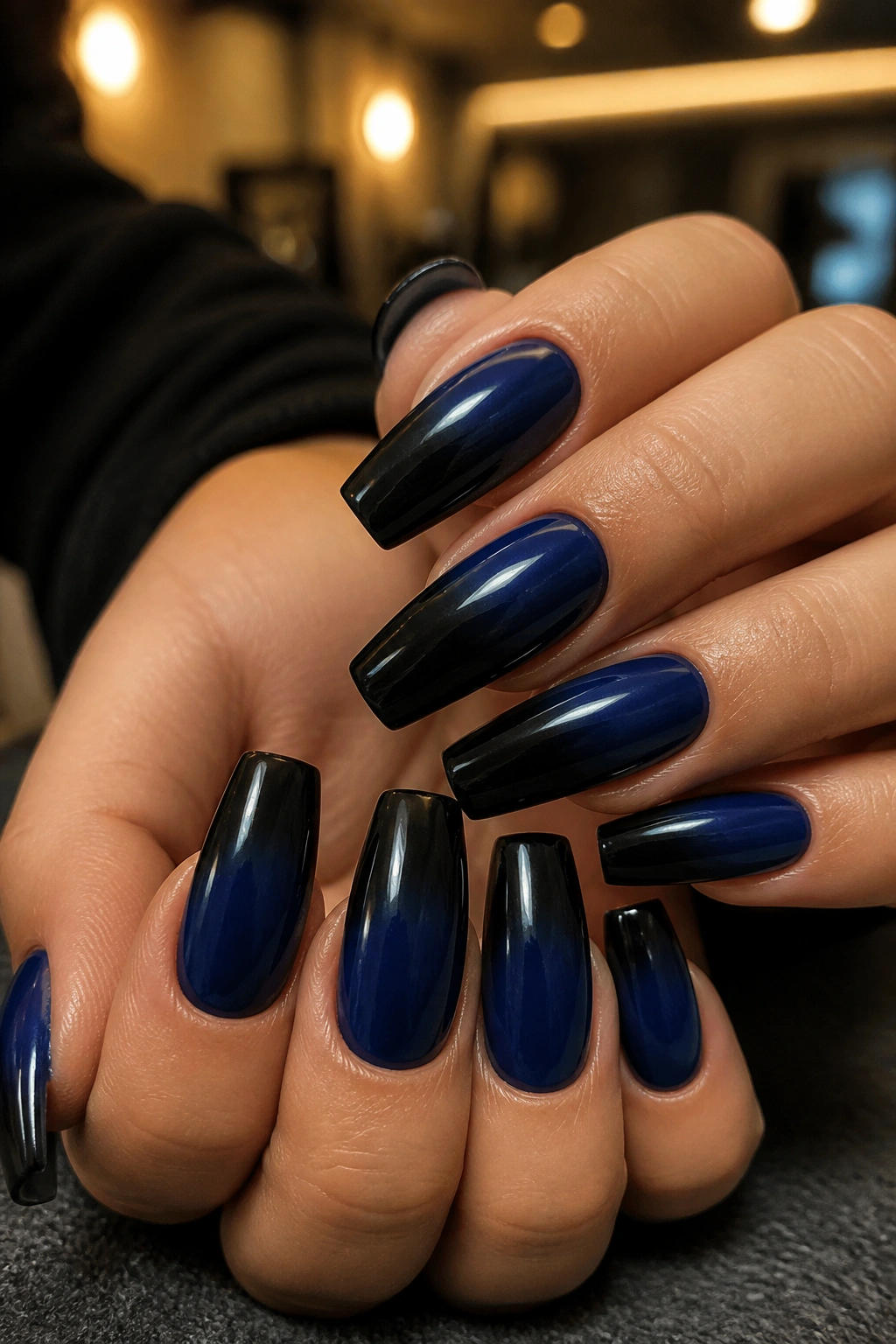 Close-up of navy-to-black ombré coffin nails on a real hand in warm salon lighting
