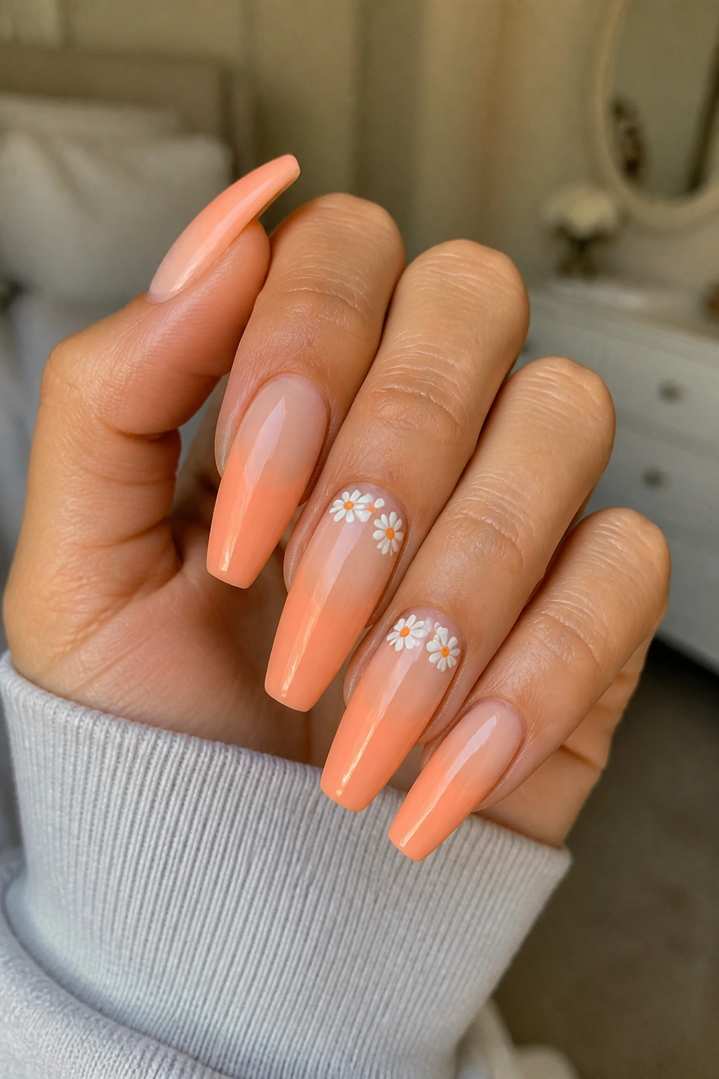 Close-up of neon peach coffin nails with tiny white daisies on a real hand in warm window-light