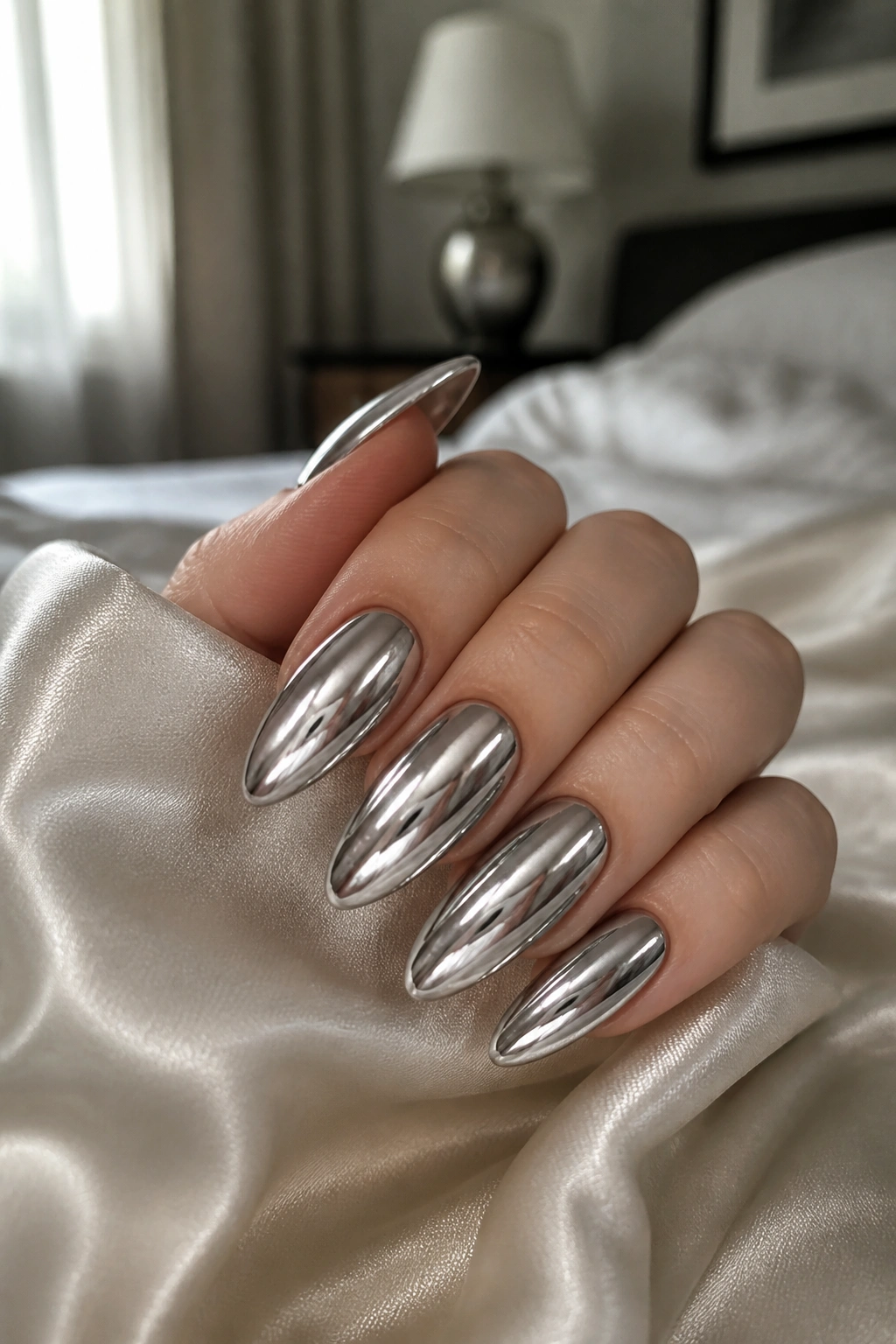 Close-up of a real hand with long almond silver nails in mirror chrome finish on satin fabric